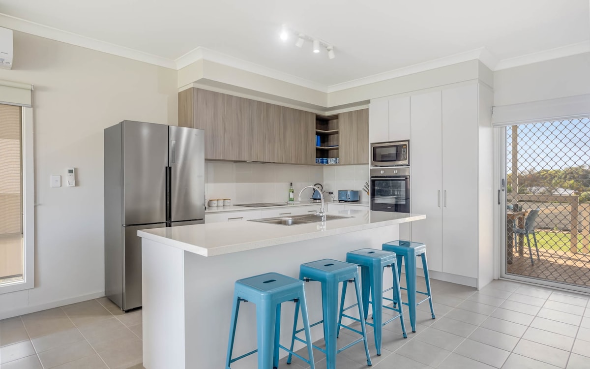 A modern kitchen features sleek cabinetry and a spacious island with four blue stools. Stainless steel appliances, including a refrigerator and ovens, complement the neutral tones. Natural light streams in through a nearby glass door, leading to an outdoor dining area.