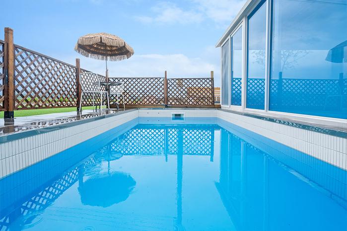 A tranquil pool area is framed by a wooden fence, featuring a sunshade and a chair nearby. The water's surface reflects the sky, creating a serene atmosphere. Large glass windows provide a view of the surrounding landscape, enhancing the sense of openness.