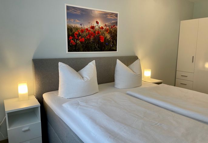 A spacious bedroom features a large bed with two decorative pillows. Nightstands with lamps are positioned on either side. A framed photograph of a poppy field adorns the wall above, bringing a touch of nature to the calming blue tones of the room.