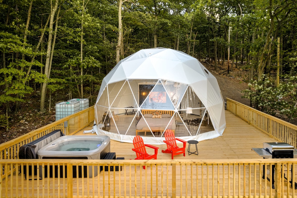 The modern glamping dome is set on a spacious wooden deck, surrounded by trees. A hot tub rests to the side, while two red chairs and a grill enhance the outdoor living space. Large geometric windows create a bright and inviting atmosphere.