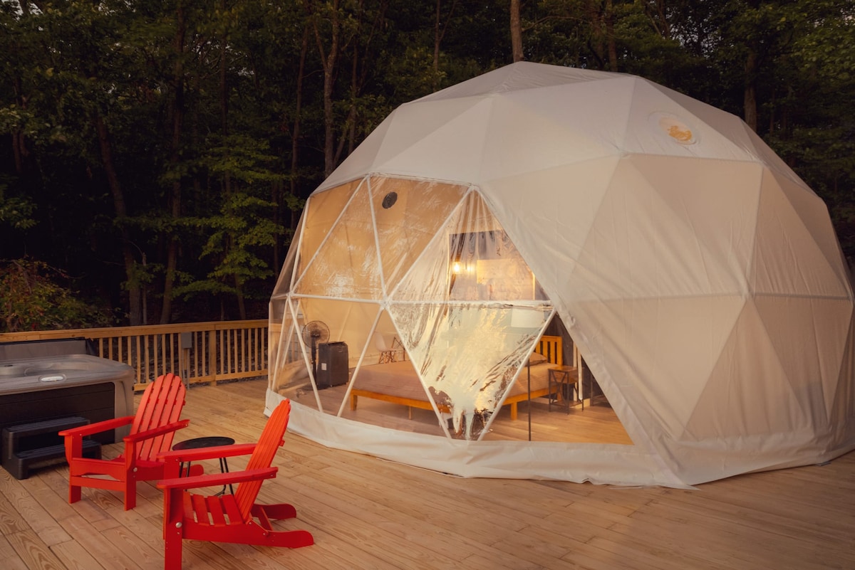 A modern glamping dome is set against a forest backdrop, featuring large transparent windows that allow for natural light. The spacious deck includes red Adirondack chairs and a hot tub, providing a cozy outdoor space for relaxation.