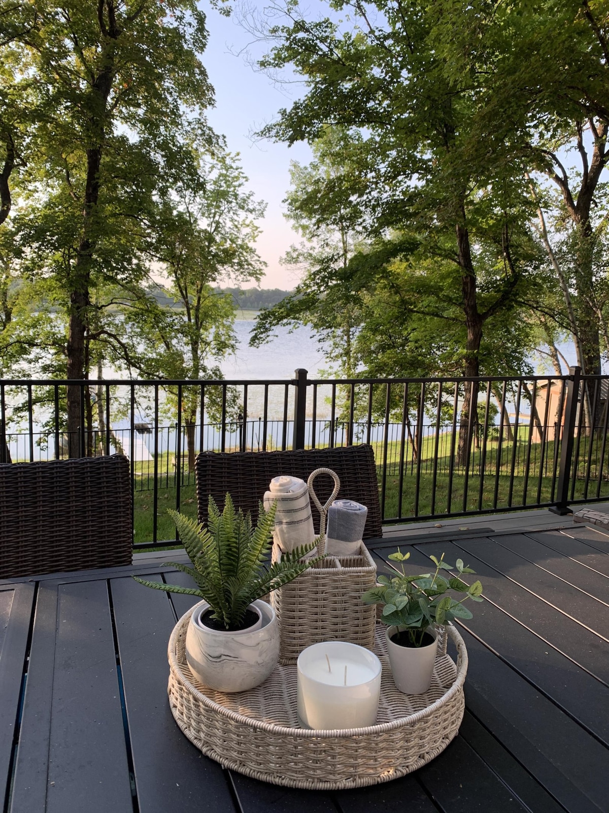 A table adorned with a woven tray sits on a wooden deck, featuring a candle, two small potted plants, and a beverage container. Green trees frame the view of McDonald Lake, enhancing the serene outdoor atmosphere.