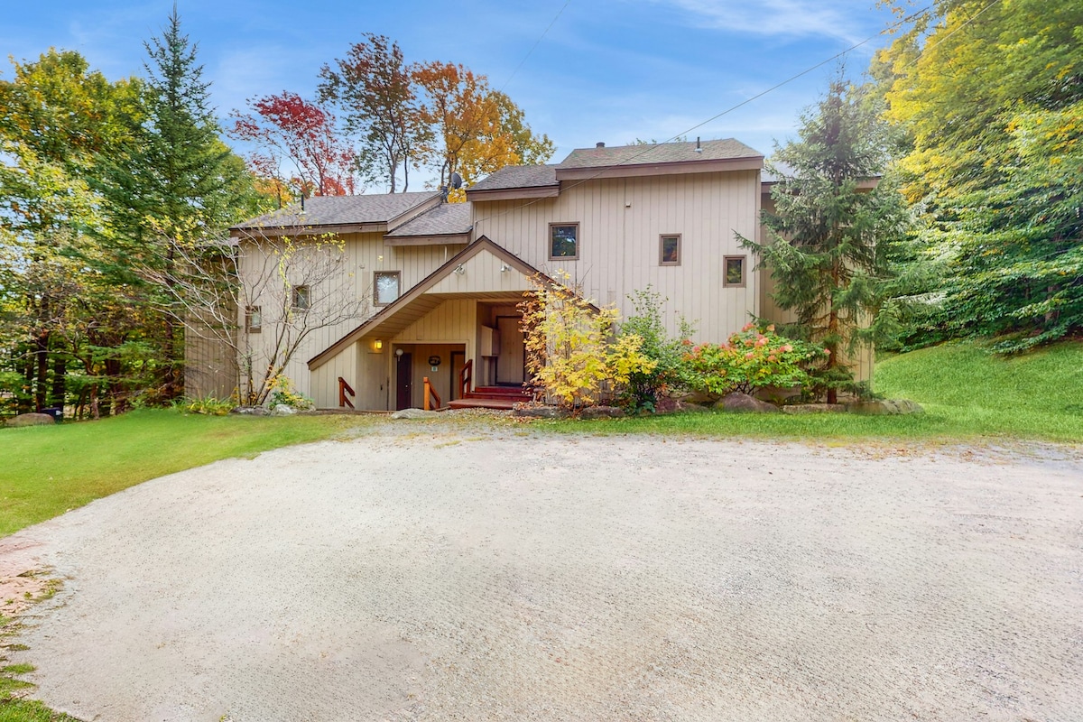 The three-level condo is nestled among trees, showcasing a welcoming entryway with steps leading up to the door. Surrounding greenery adds to the natural feel, while a gravel area provides parking for several vehicles.