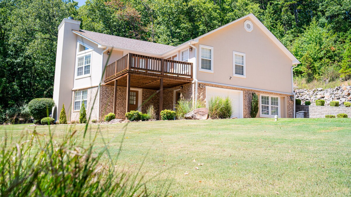 The exterior of the property showcases a two-story home surrounded by landscaped greenery. A spacious deck is visible on the upper level, while the lower level features a combination of brick and siding. The setting is serene, with tree-covered hills in the background.
