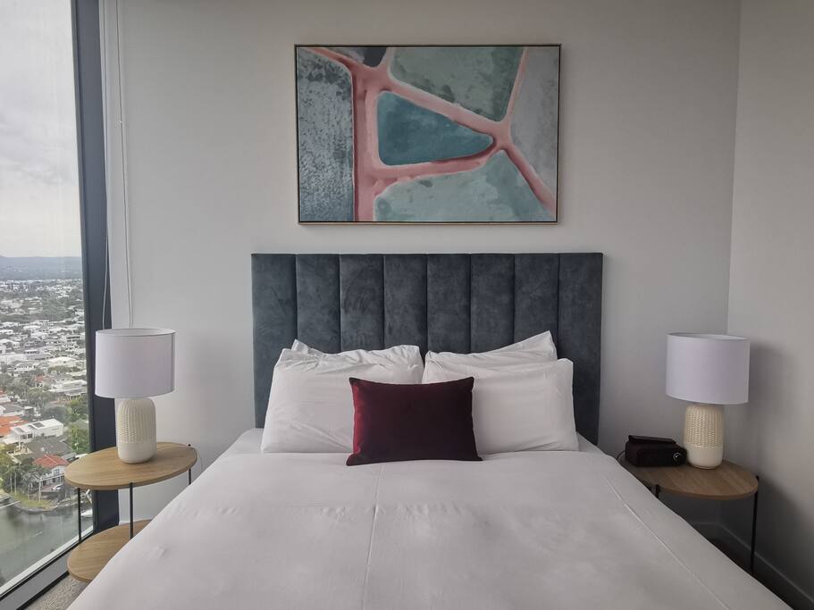 A sleeping area features a spacious bed with plush white linens and a burgundy accent pillow. Flanking the bed are two round side tables with soft glow lamps. A wall art piece in soft hues adds a contemporary touch, with large windows providing scenic views.