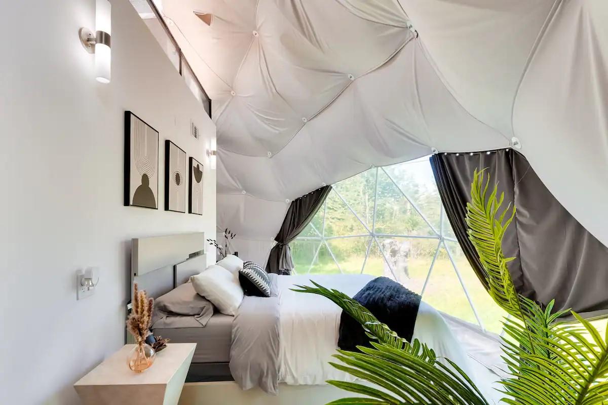 A spacious sleeping area features a cozy bed adorned with soft linens. A geometric wall design enhances the modern aesthetic, while large windows invite natural light and offer views of the lush exterior. Decorative plants add a touch of greenery to the serene environment.