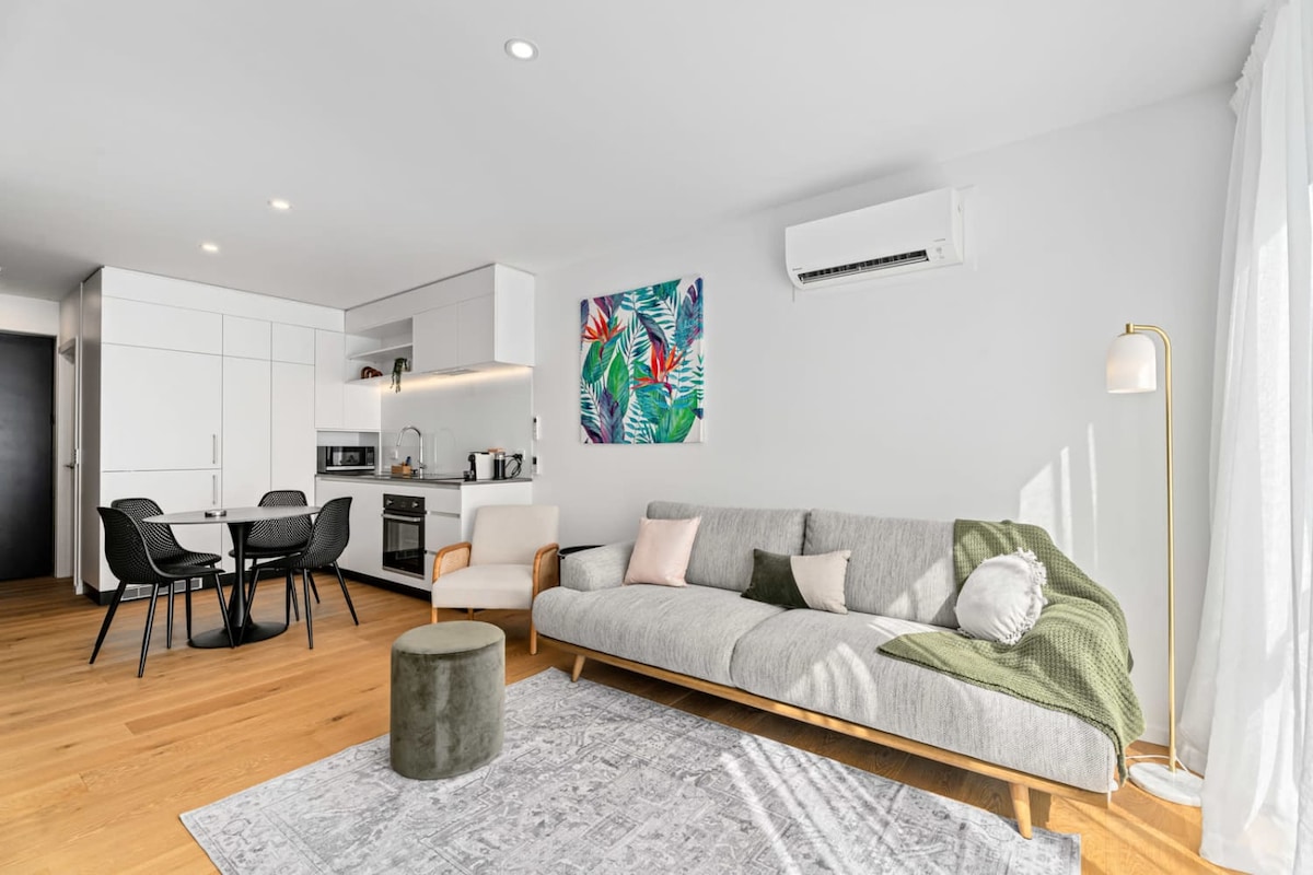 A modern living area features a light grey sectional sofa with accent pillows and a green throw, complemented by a round, upholstered ottoman. A sleek dining table with four chairs is visible, along with a well-equipped kitchen area. Natural light streams in through sheer curtains, enhancing the space.