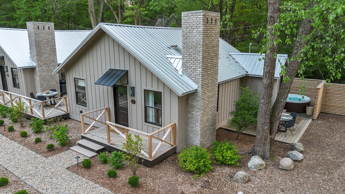 The exterior of a cottage is shown, featuring a light-colored wooden façade and a metal roof. A welcoming entryway and landscaped pathway are visible, along with stacked stone chimneys. A private deck area, including a hot tub and outdoor seating, is situated in the background among greenery.