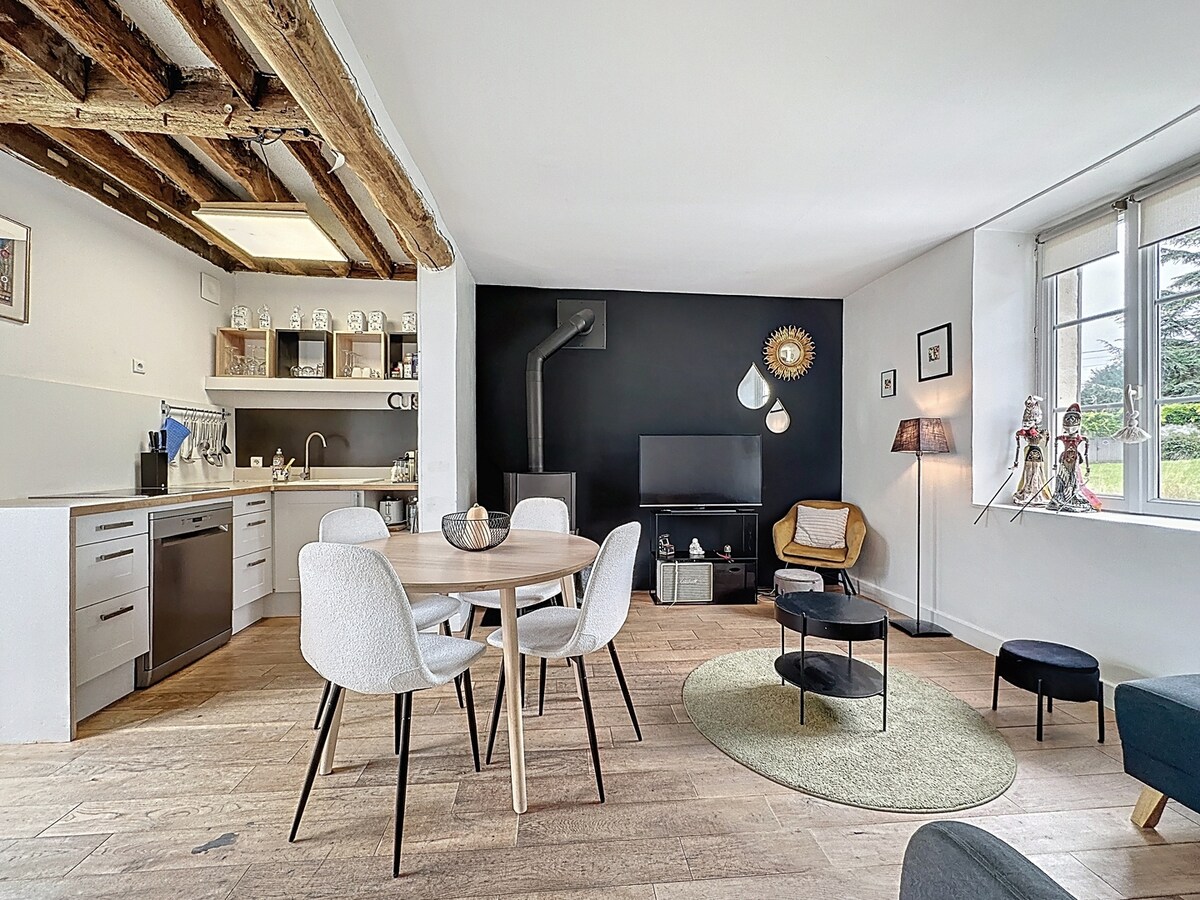 An open and airy living space features a dining table surrounded by light-colored chairs. A sleek kitchen area is visible, along with a cozy seating arrangement. A flat-screen TV rests on a black accent wall, complemented by various light fixtures and decorative elements.