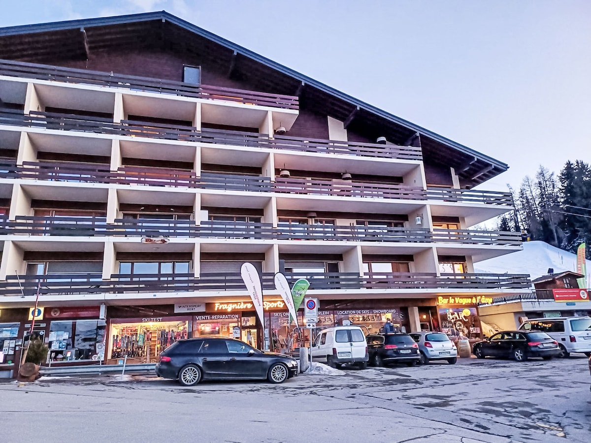 A four-story residence is seen from the street, featuring a wooden facade with balconies. Businesses occupy the lower level, with signage for a sports shop and a bar. Various parked cars are visible in front of the building, set against a backdrop of trees and mountains.