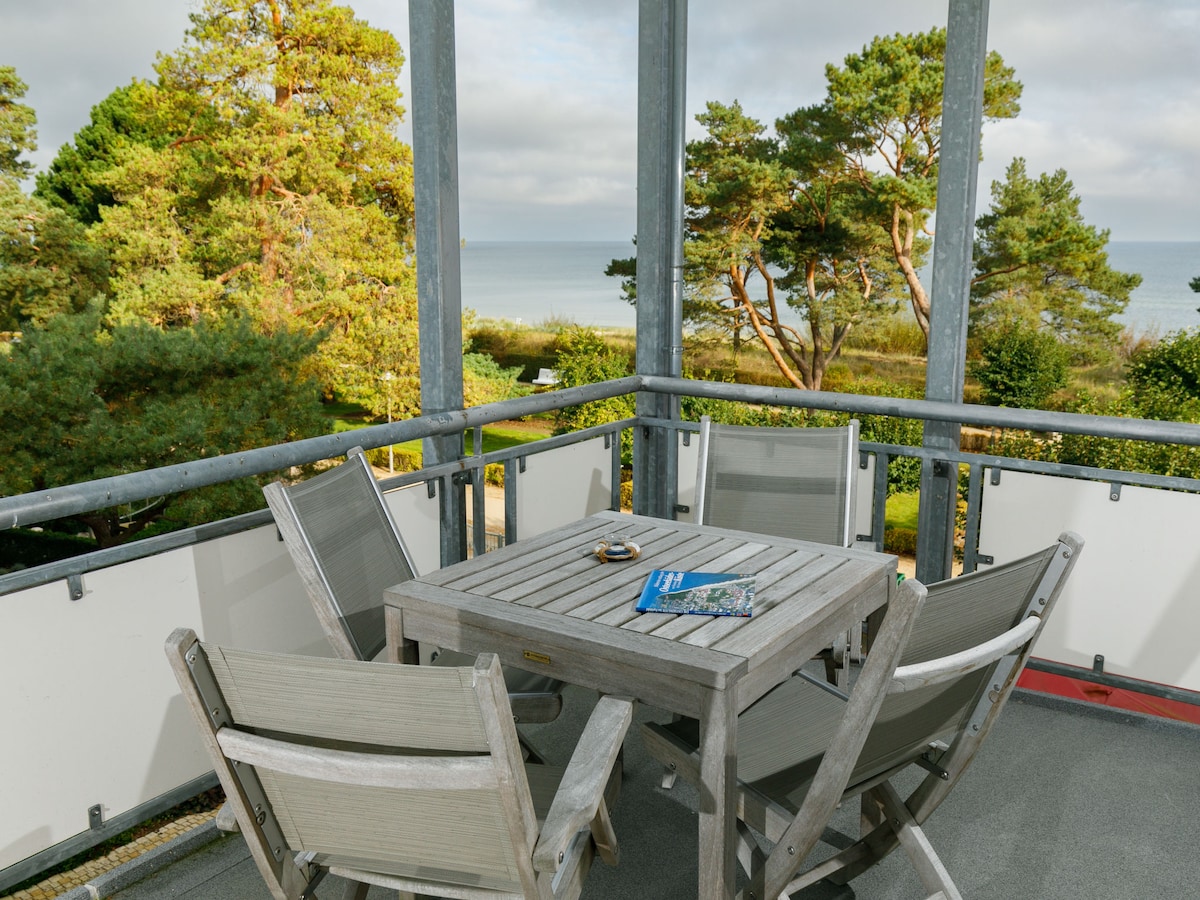 A balcony space is furnished with a wooden table and four chairs, offering a view of the sea and surrounding greenery. Light clouds hover above, and the tranquil setting features a serene coastal atmosphere.