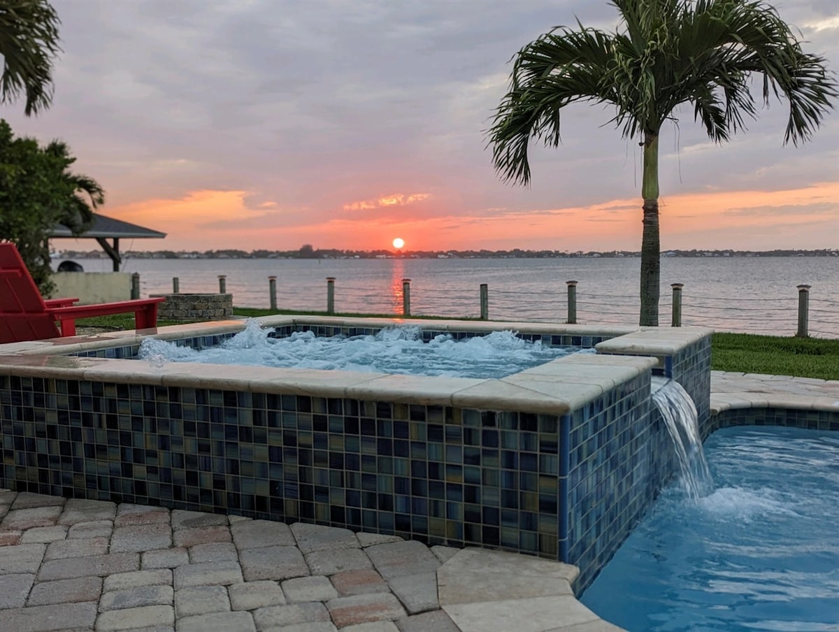 A hot tub with cascading water features is situated beside a tranquil river. The sunset casts warm hues across the sky, while palm trees frame the scene, providing a tropical ambiance. Paved stones surround the hot tub, enhancing the outdoor setting.