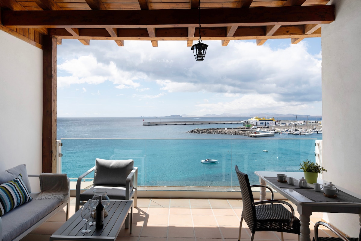 A private covered terrace showcases stunning views of the Atlantic Ocean. Comfortable seating is arranged around a low table, while glass railings enhance the panoramic vista. The peaceful scene includes boats resting on the calm water, complemented by a gentle sky and distant shoreline.