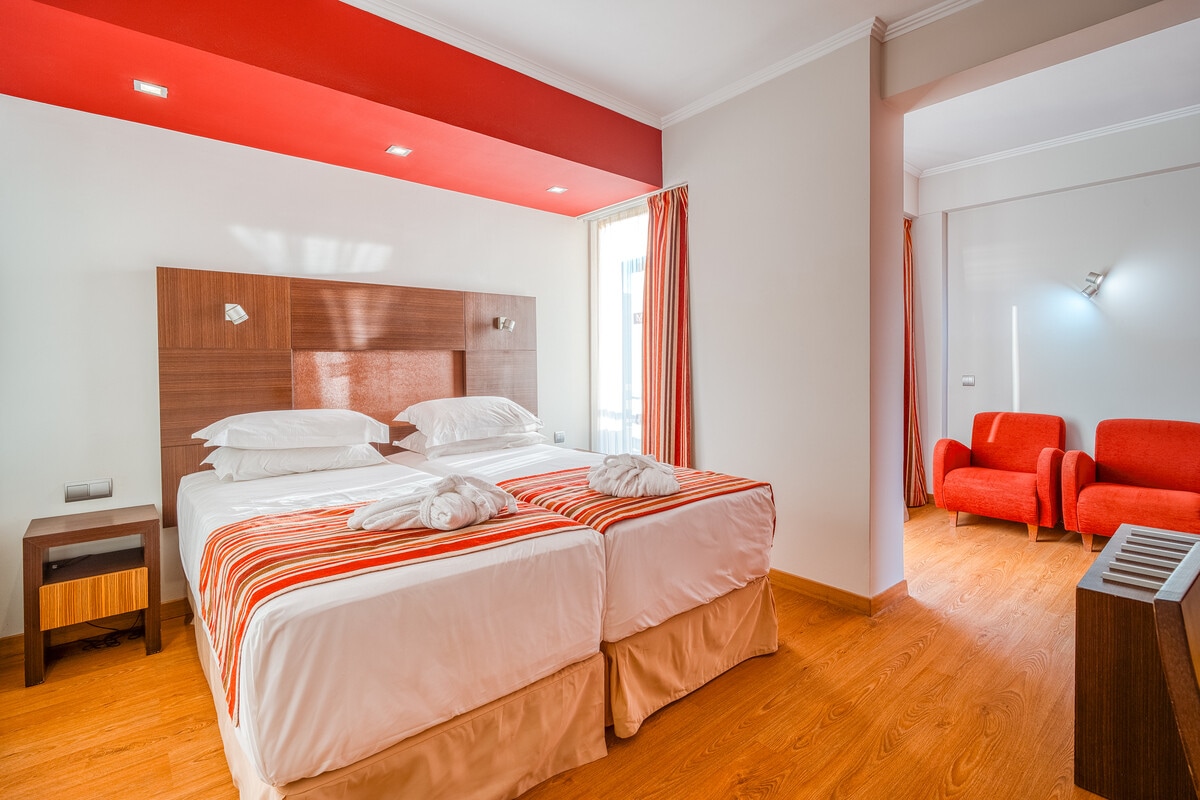 A well-lit room features a double bed with a light-colored headboard and crisp white linens, complemented by vibrant red accent walls. Two armchairs in matching red are positioned near the window, enhancing the overall spaciousness of the room.
