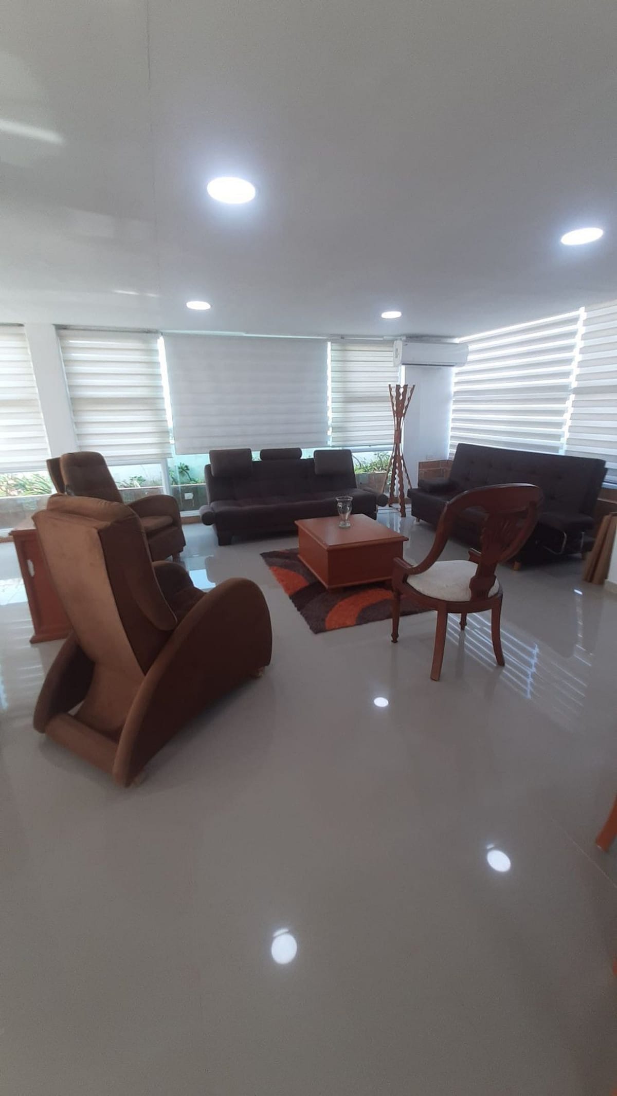 A bright living area features several comfortable seating options, including two armchairs and a two-seater sofa. A wooden coffee table is centered on a decorative rug, while large windows allow natural light to fill the space, complemented by soft window treatments.
