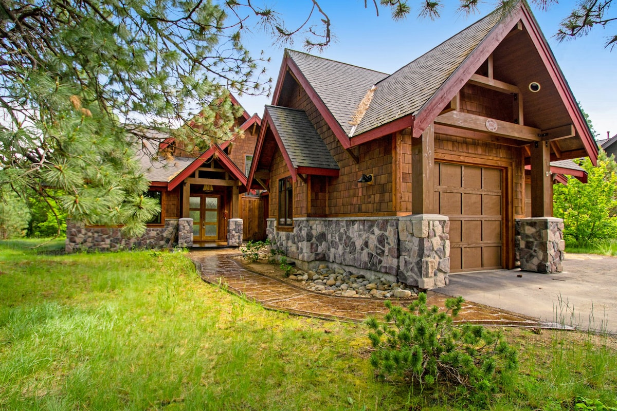 The exterior of the home features a combination of wood and stone materials, framed by lush greenery. A charming entryway with a covered porch welcomes visitors. The garage door is visible, blending with the natural environment surrounded by manicured landscaping.