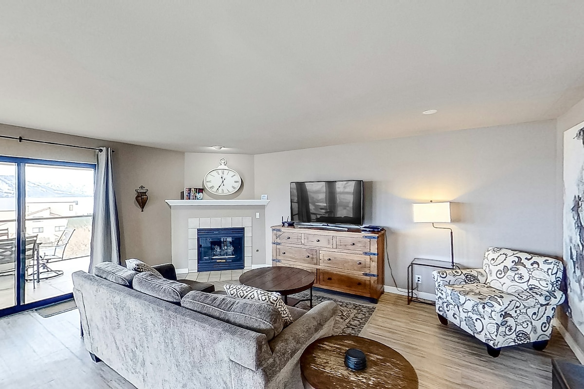 A cozy living area is presented with a plush sofa and a decorative armchair. A large TV is mounted above a wooden cabinet, and a fireplace adds warmth. Natural light streams in through sliding glass doors that lead to a patio, enhancing the inviting space.