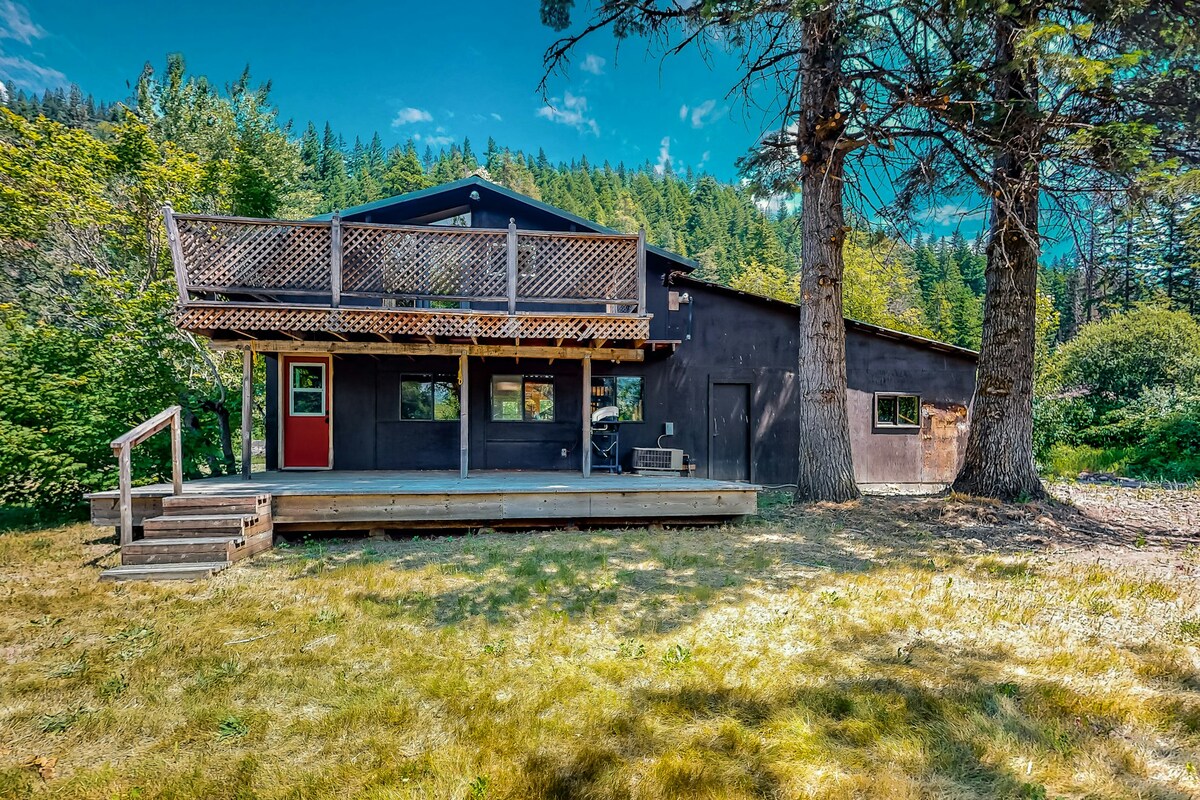 The exterior of a two-story retreat is shown, featuring a wooden deck with stairs leading to the entrance. Large windows offer views of the surrounding greenery, and a red door adds a contrasting element. Towering trees provide shade, enhancing the natural setting.