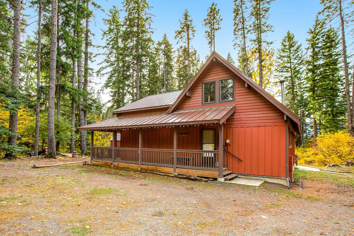 A charming red house is nestled among tall trees in a tranquil forest setting. The exterior features a covered porch with railings, providing a welcoming entryway. The surrounding area includes a gravel pathway and foliage, highlighting the natural beauty of the location.