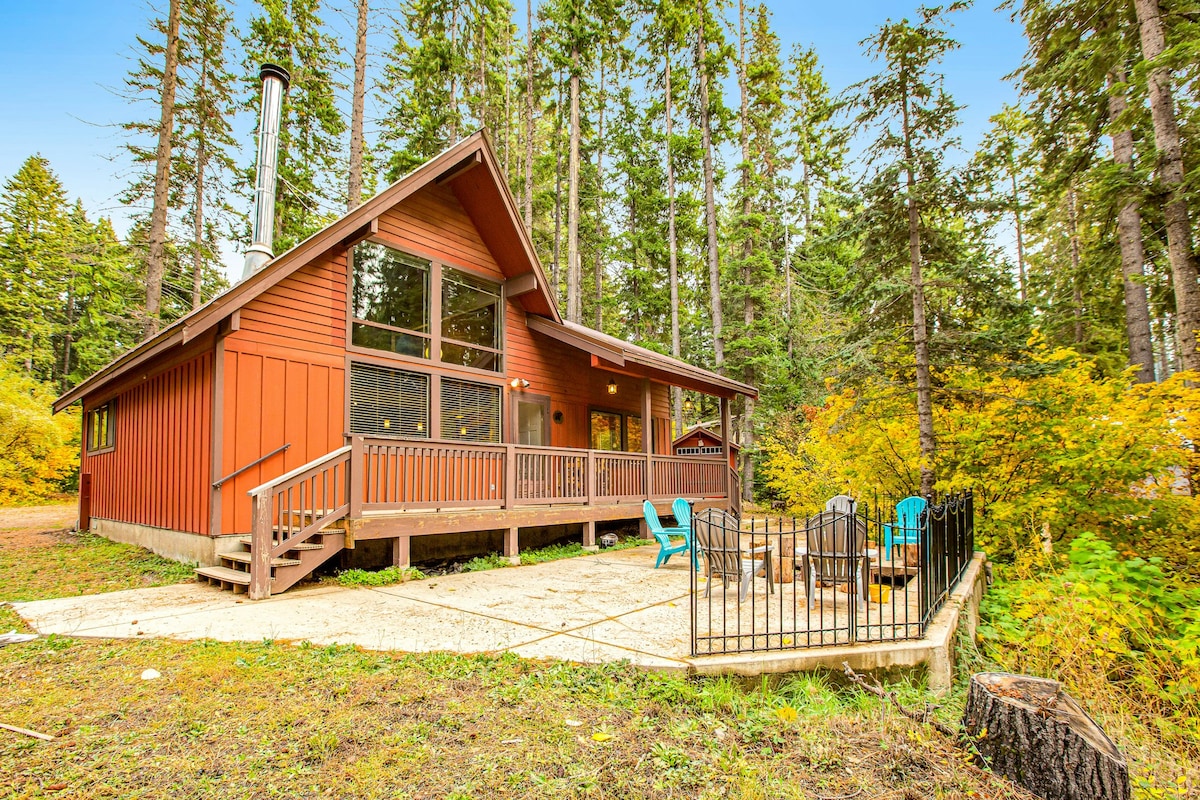 The charming lakehouse is set against a backdrop of tall trees, featuring a wraparound deck with colorful outdoor seating. Large windows allow ample natural light, while the outdoor firepit area invites cozy gatherings in the peaceful forest setting.