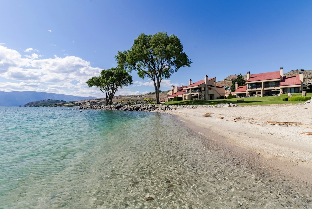 A serene view of a sandy beach with clear water leading to the lake. Behind, modern condo units are visible, framed by grass and trees under a bright sky. The landscape invites relaxation and outdoor activities.