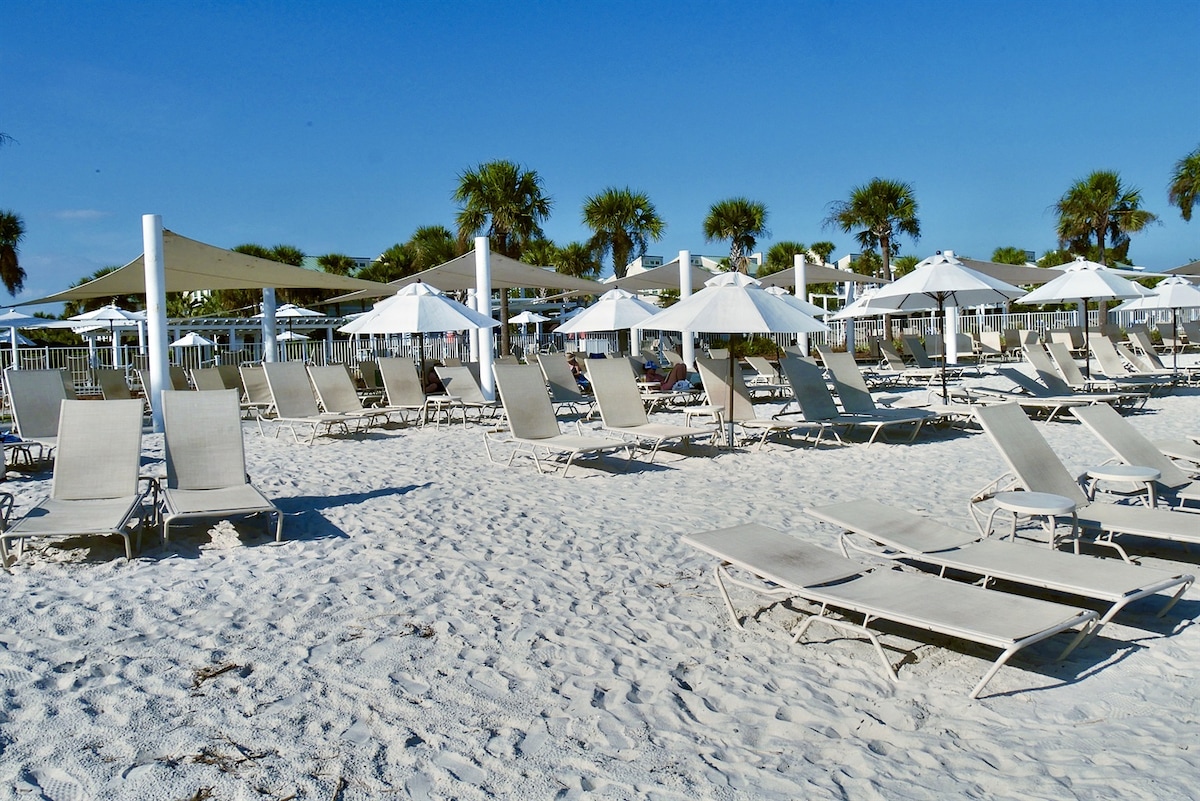 A spacious beach area is filled with lounge chairs arranged on soft white sand. Large umbrellas provide shade overhead, while palm trees dot the background, creating a relaxed coastal atmosphere.