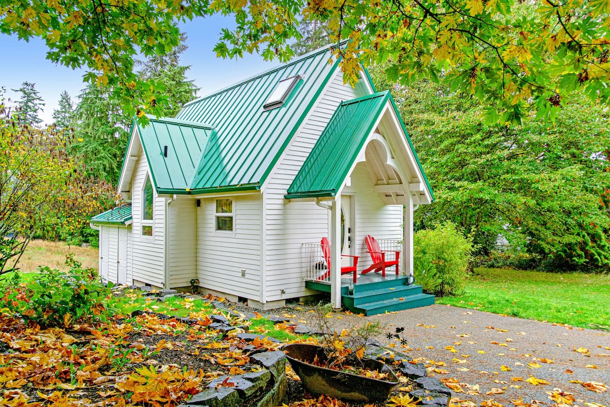 The cottage features a distinctive white exterior accented by a green metal roof. A welcoming porch is furnished with two red Adirondack chairs. Surrounding foliage showcases autumn leaves, adding a warm touch to the vibrant natural landscape.