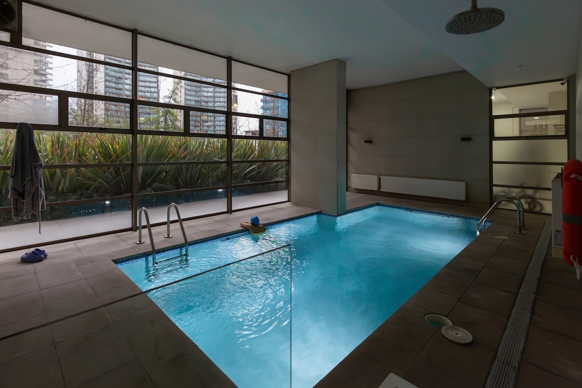 An indoor swimming pool is visible, surrounded by large glass windows that allow natural light to illuminate the water. A few steps lead into the pool, and seating areas are located beside it. Lush greenery can be seen outside through the windows.