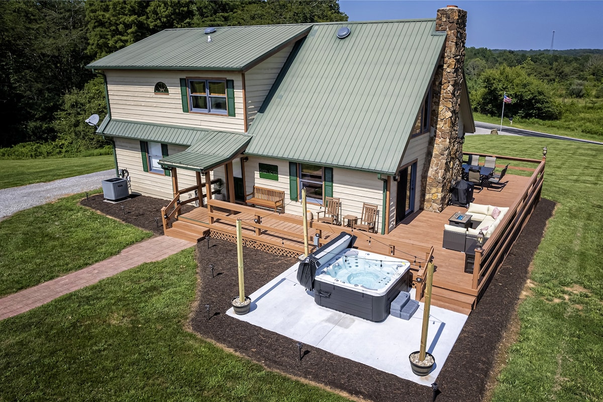 An exterior view of the country chateau showcases a welcoming deck with seating, surrounded by lush greenery. A private hot tub is positioned on a concrete pad, adjacent to the home, enhanced by a stone chimney that adds rustic charm.