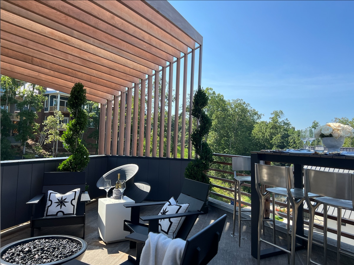 A rooftop terrace showcases comfortable seating arrangements alongside a high-top dining table. Greenery is thoughtfully incorporated, enhancing the space's natural appeal. Clear blue skies provide a serene backdrop, while the terrace offers a stylish setting for relaxation and gatherings.