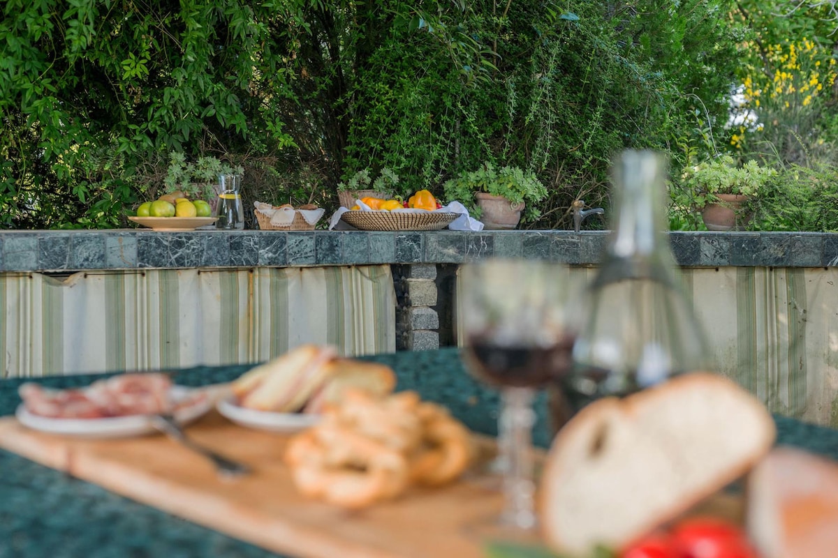 An outdoor dining area is set against a backdrop of lush greenery. A marble countertop holds an array of fresh fruits and a selection of dishes, with a wooden cutting board displaying sliced bread and snacks in the foreground. Glassware and utensils complement the inviting scene.