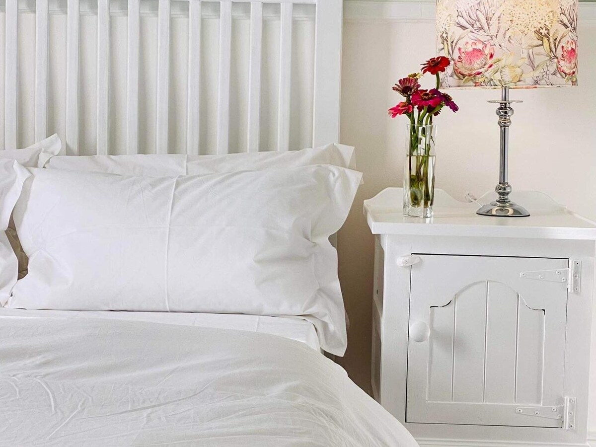 A bright sleeping area is presented with a spacious bed dressed in crisp white linens. A bedside table features a glass vase with fresh flowers and a decorative lamp, adding a touch of color to the muted tones of the room.