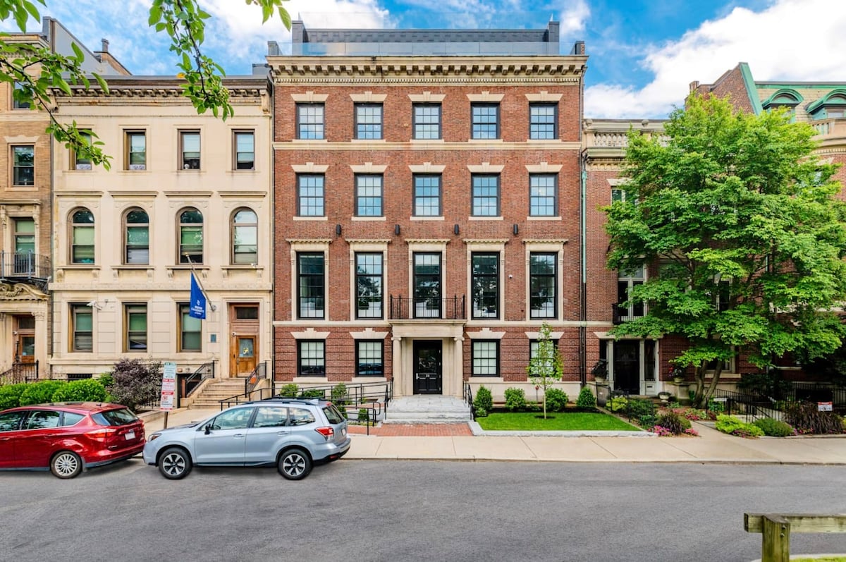 The exterior of a historic brick building features a grand entrance with marble steps and a well-maintained facade. Large windows on each floor provide a view of the street lined with trees and parked cars. The surrounding area showcases tasteful landscaping.