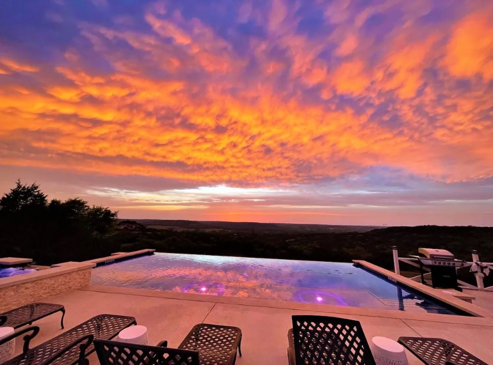 A stunning sunset paints the sky in vibrant hues above a serene infinity pool. The pool's edge seamlessly reflects the colorful clouds, while several lounge chairs are positioned nearby, offering a perfect spot to enjoy the breathtaking hill country views.