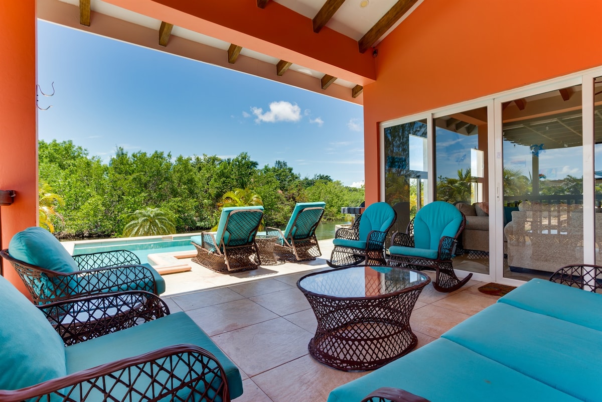 An outdoor lounge area features turquoise seating and a glass-top table, set against a backdrop of lush greenery and a private pool. Sun loungers are arranged for relaxation under the sun, while large sliding doors provide access to the interior living space.