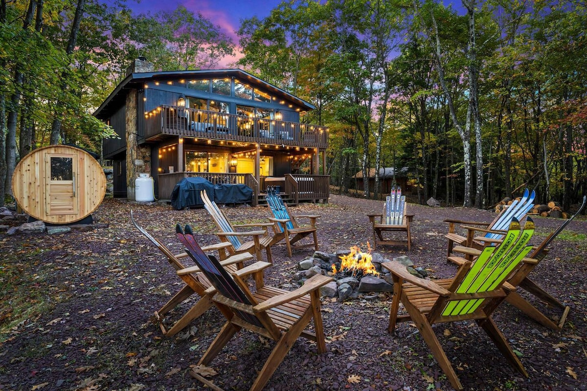 A cozy outdoor fire pit area is surrounded by colorful adirondack chairs, inviting relaxation and conversation. The backdrop features a spacious cabin with large windows, enhancing the connection to nature as twilight casts a soft glow over the landscape.