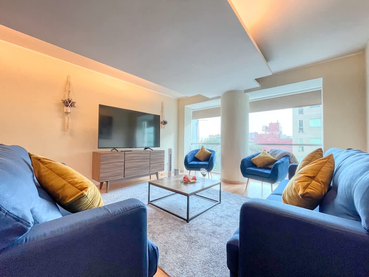 A welcoming living area features two blue sofas and two accent chairs with yellow cushions. A contemporary coffee table sits at the center, while a large television is mounted on the wall. Natural light fills the space through expansive windows, enhancing the neutral tones and modern decor.