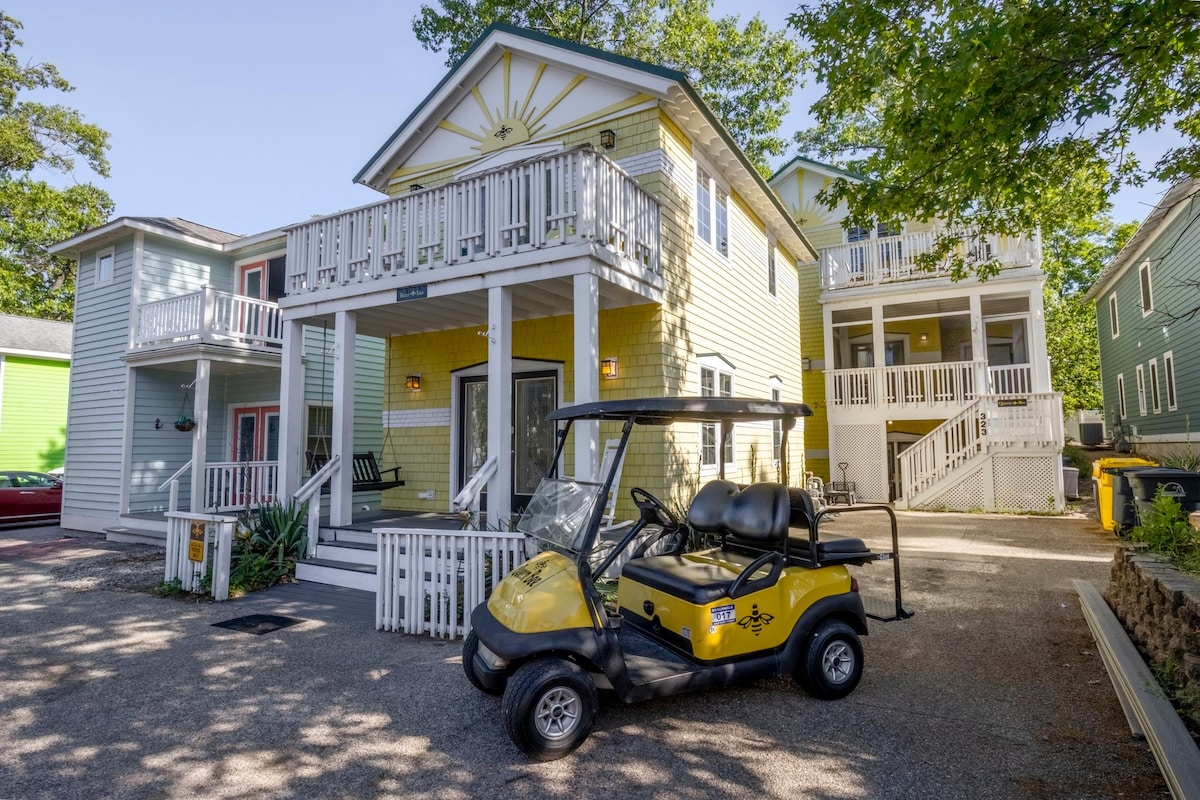 A vibrant yellow house with a decorated porch stands prominently, flanked by greenery and additional colorful homes. A golf cart with rear seats is positioned in front of the house, providing convenient transportation for guests.