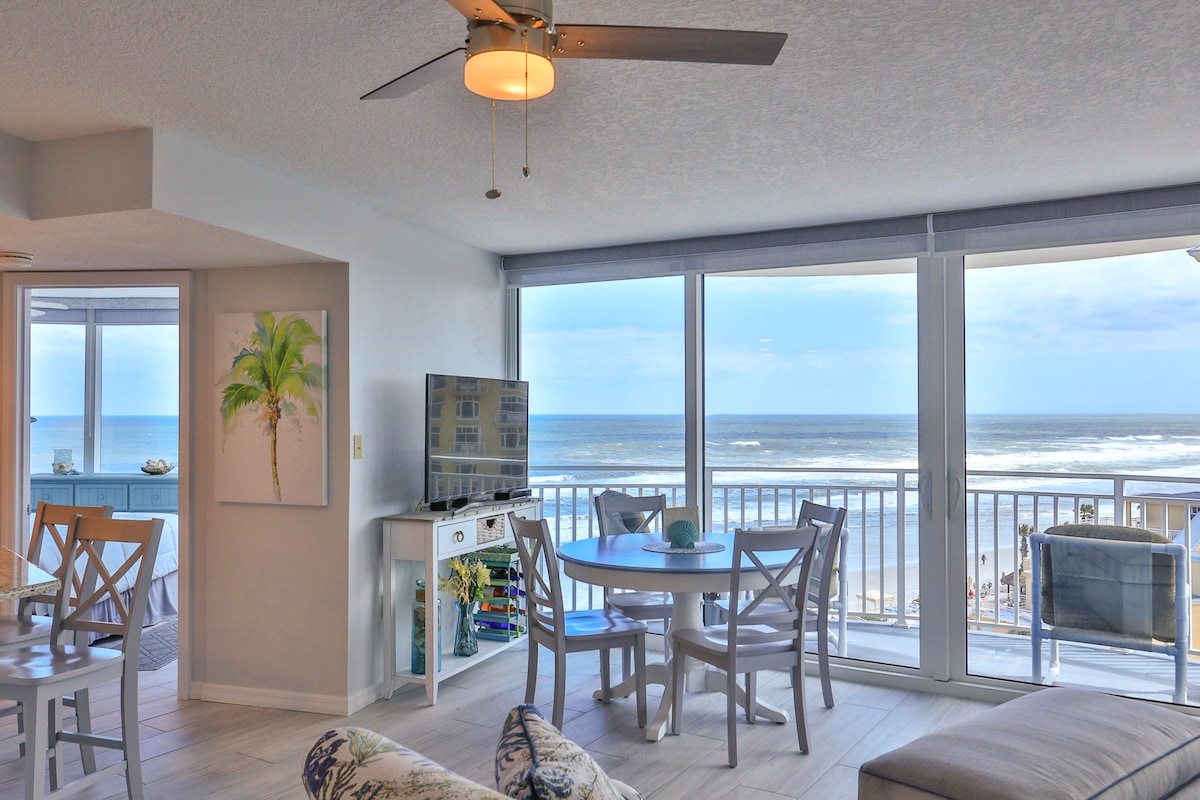 A spacious living and dining area features large sliding glass doors that open to an oceanfront balcony. A round dining table with chairs is positioned near the window, while a comfortable seating area includes a patterned sofa. A ceiling fan enhances the airy atmosphere.