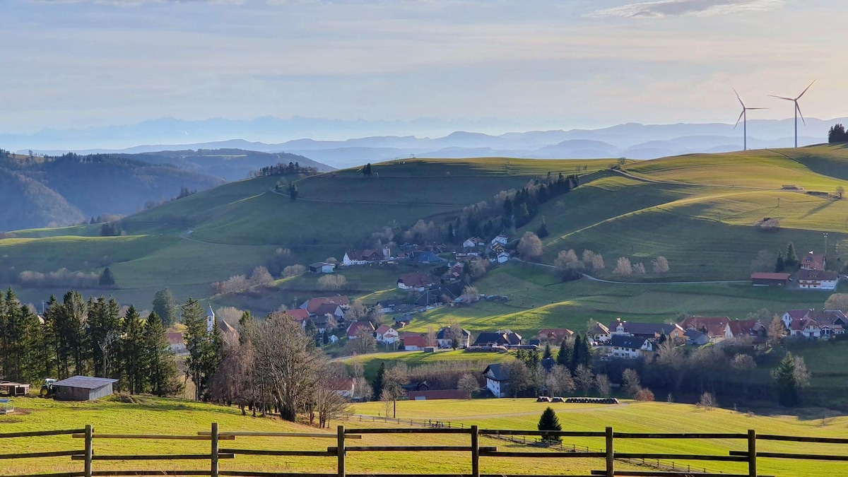 A scenic view of rolling green hills is framed by a wooden fence, showcasing a small village nestled among the natural landscape. In the distance, wind turbines stand against a hazy sky, complementing the serene countryside setting.