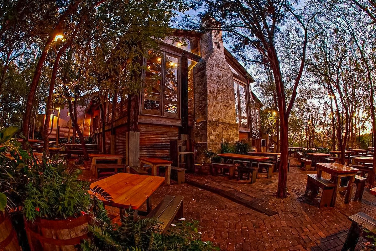 An outdoor dining area features rustic wooden tables and benches, surrounded by lush greenery and trees. The stone building creates a cozy ambiance, while warm lights illuminate the space, enhancing the inviting atmosphere during dusk.