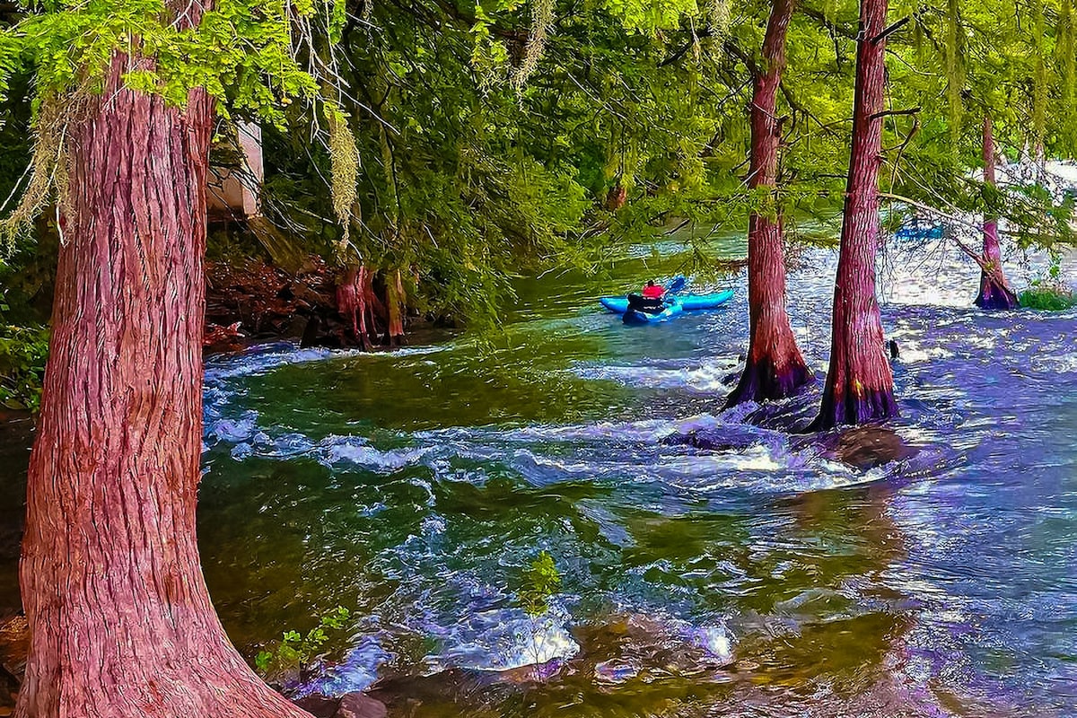 Kayakers navigate the gentle waters of the Guadalupe River, flanked by tall cypress trees. Sunlight glimmers on the water's surface, creating a serene atmosphere. The lush greenery and textured tree trunks enhance the natural beauty of this riverside setting.