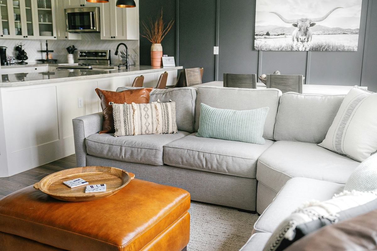 A soft sectional sofa in neutral tones is accompanied by decorative pillows in various textures. A wooden tray with coasters sits atop a leather ottoman. The kitchen area with light cabinetry and stainless steel appliances is visible in the background, adding a modern touch.