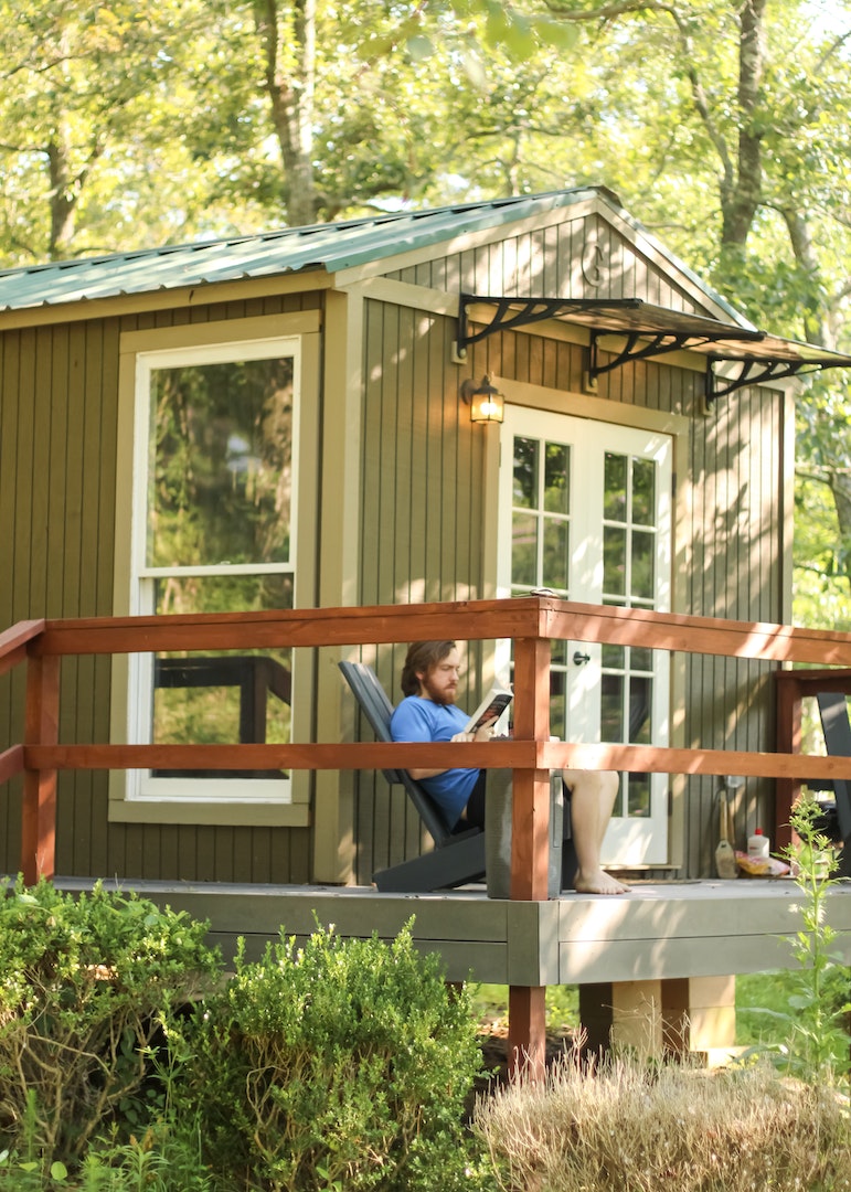 A quaint tiny house is nestled among trees, featuring a wrap-around porch with a comfortable chair. Large windows invite natural light, while the rustic finish complements the serene surroundings. The porch railing adds an inviting touch to the outdoor space.