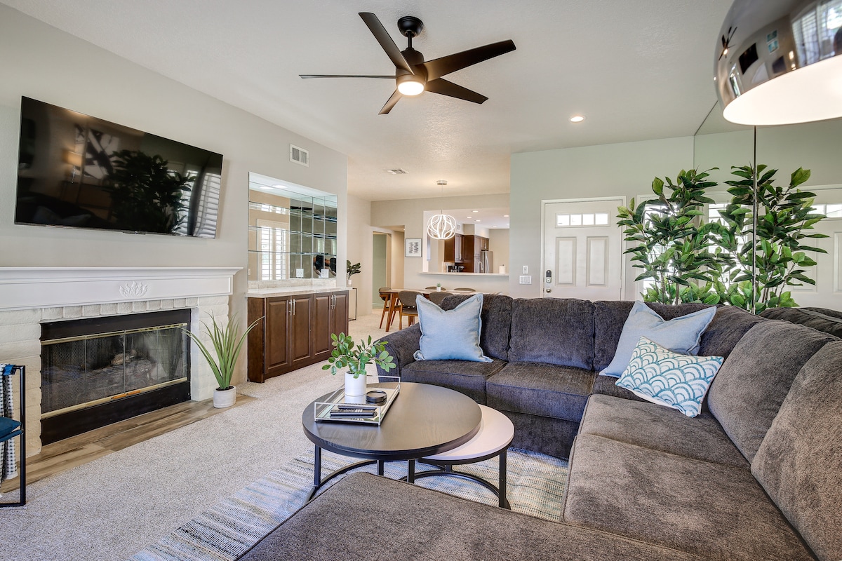A spacious living area is featured, showcasing a large sectional sofa in neutral tones with accent pillows. A flat-screen television is mounted above a gas fireplace. A round coffee table sits in the center, surrounded by greenery and soft carpet, creating a comfortable gathering space.