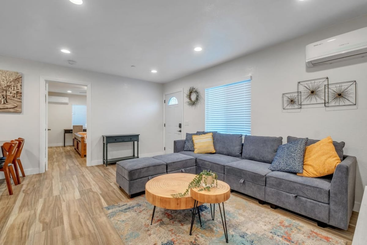 A spacious living area features a large sectional sofa with colorful throw pillows, complemented by a circular wooden coffee table. Light-colored walls and modern lighting create a bright setting. A doorway leads to additional rooms, and a window allows natural light to fill the space.