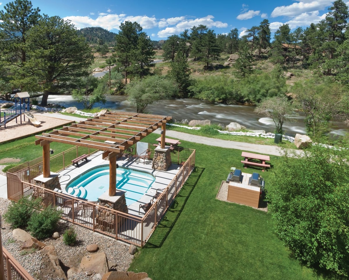 An outdoor area features a seasonal pool surrounded by stone and a wood pergola. Nearby, a picnic area with tables overlooks a flowing river, with trees and green grass enhancing the natural setting.
