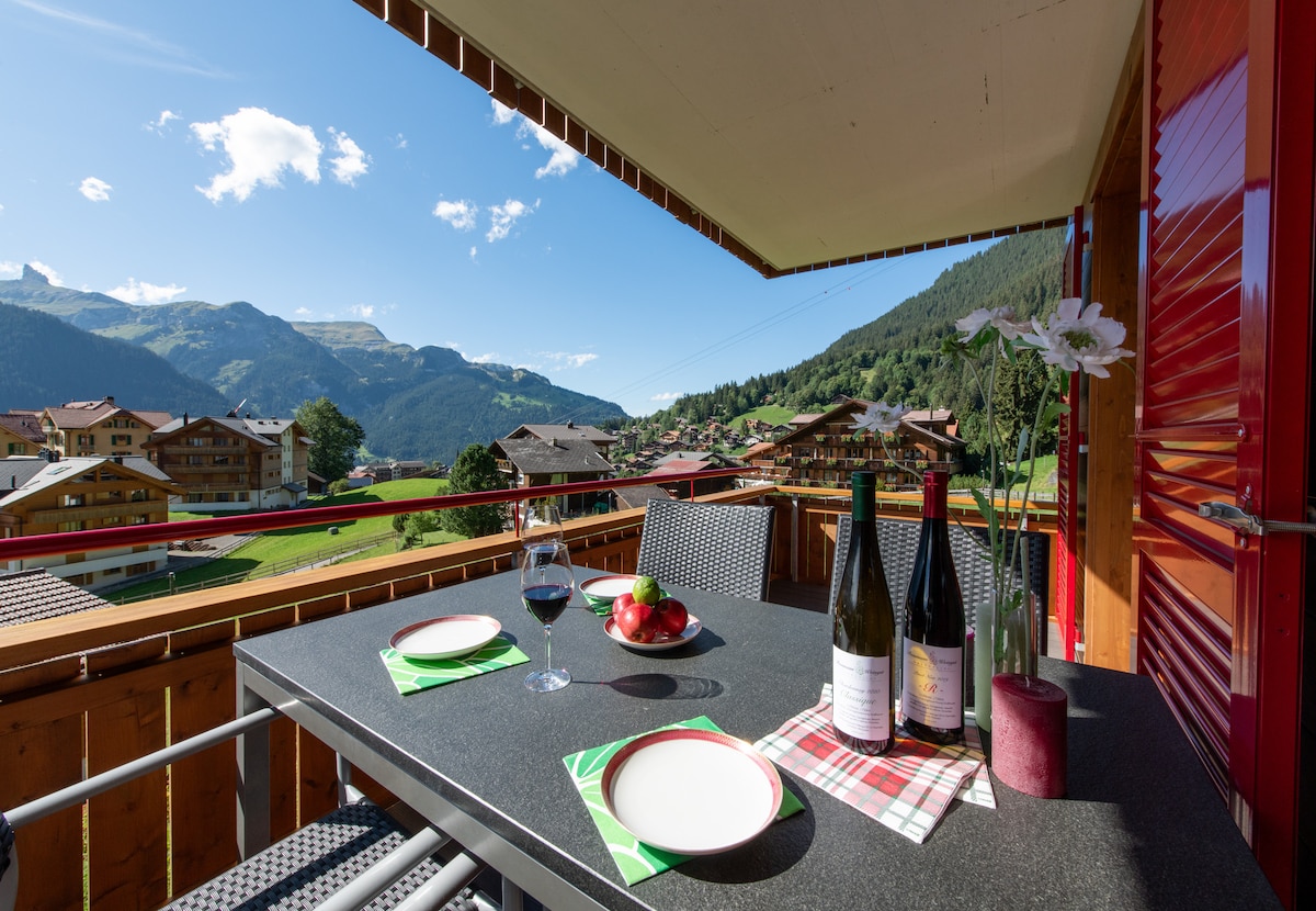 A west-facing balcony features a dining table set for four, with plates and glasses of wine. Scenic mountain views are visible in the background, framed by sunny skies and scattered clouds. The surrounding area showcases charming village architecture.