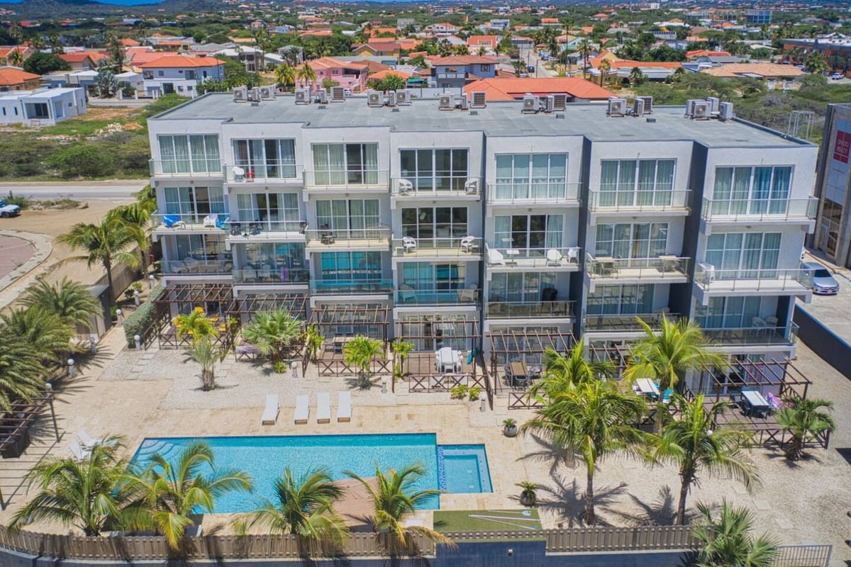The exterior of the three-story condo complex is depicted, showcasing a modern design with large glass windows. Surrounding palm trees and a spacious shared pool are visible at the forefront, while the neighborhood features colorful rooftops in the background.