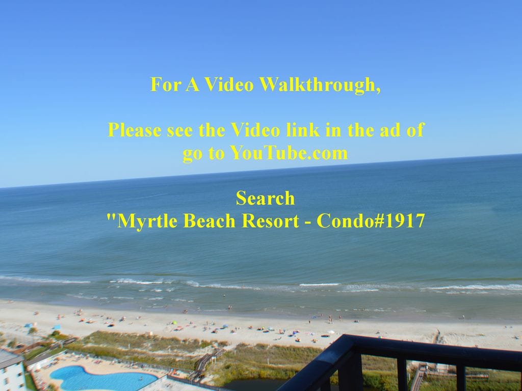 An ocean view is showcased from a balcony, featuring expansive blue waters and a sandy beach below. The coastal landscape is framed by a clear sky, creating a serene atmosphere. Beachgoers can be seen enjoying the shoreline, with a pool visible in the foreground.